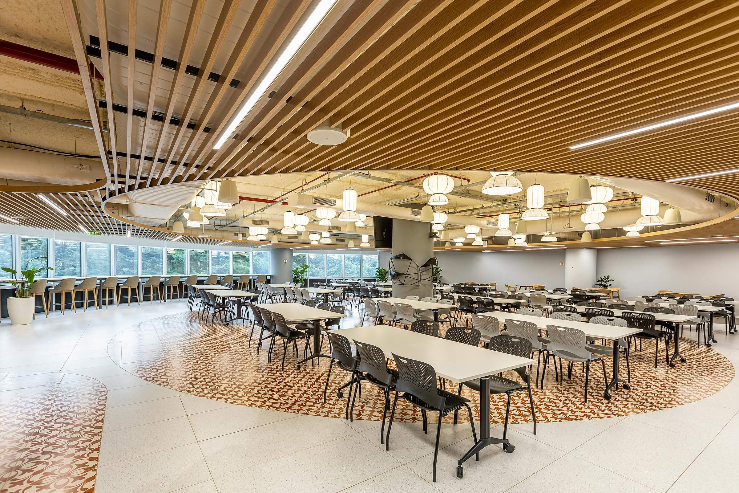 Large cafeteria with circular ceiling feature, long tables, mixed seating, and perimeter windows