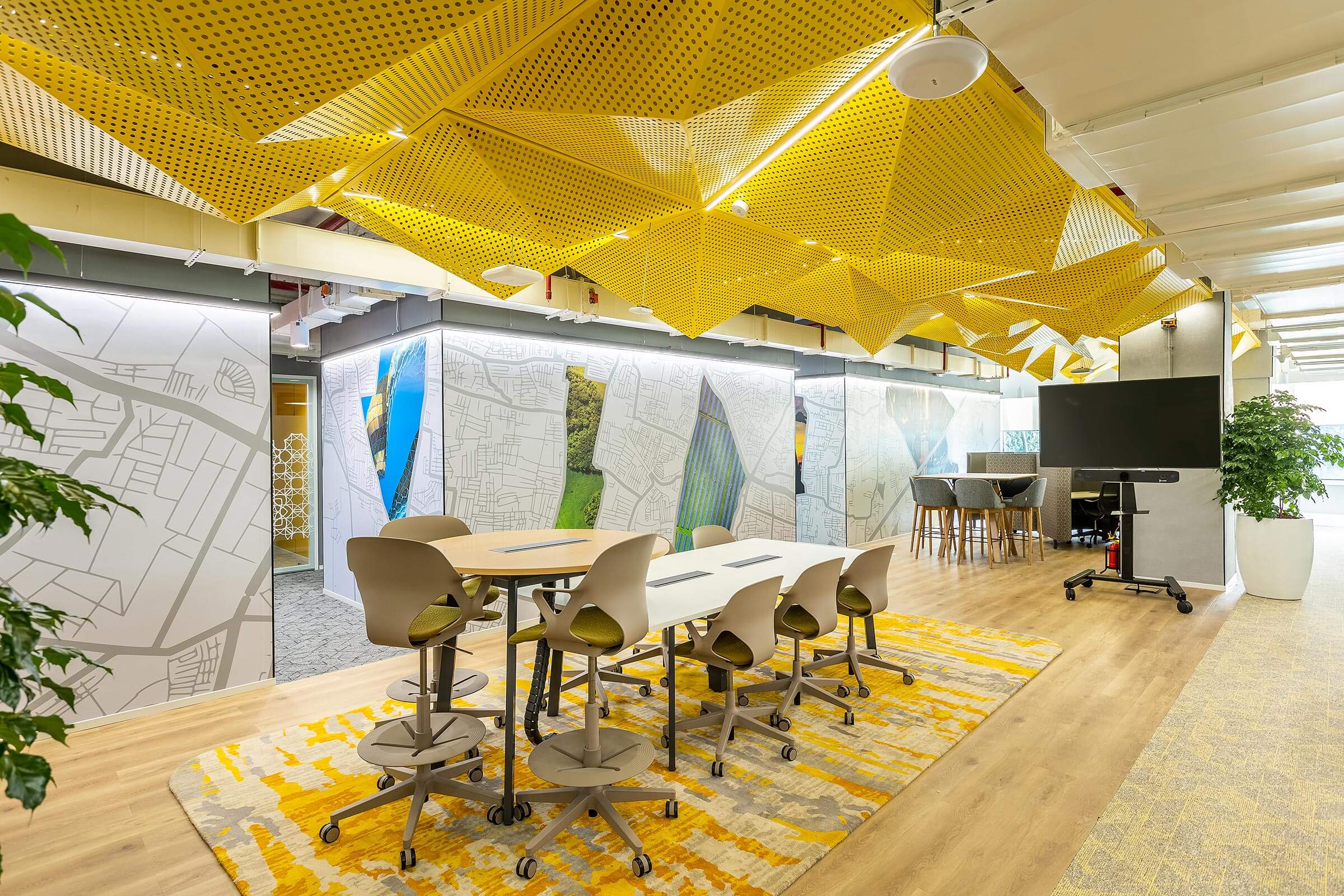 Open workstation area with high tables, mobile chairs, yellow ceiling panels, and collaborative screens