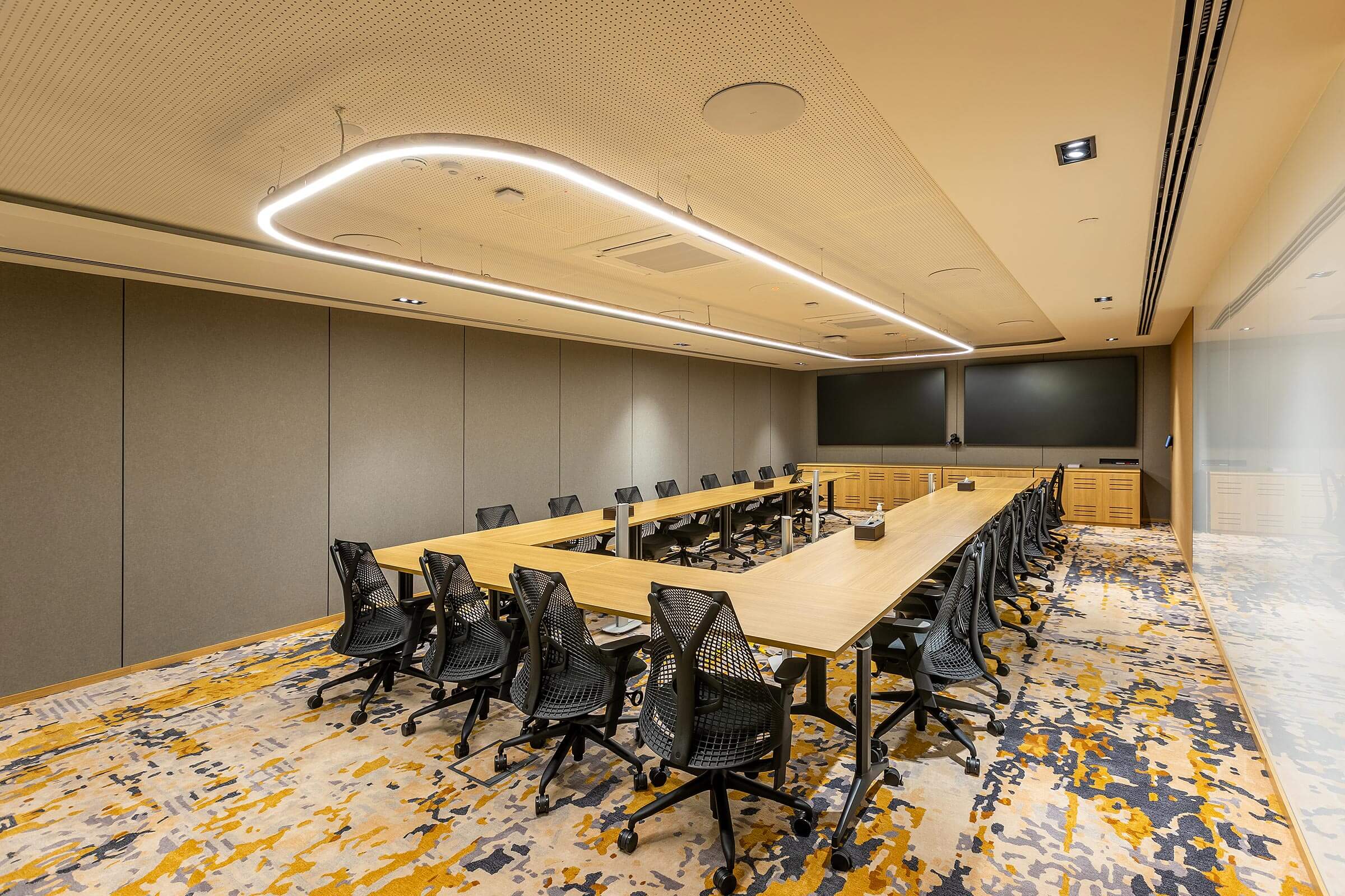 Long conference table with task chairs, dual display screens, and patterned carpet flooring