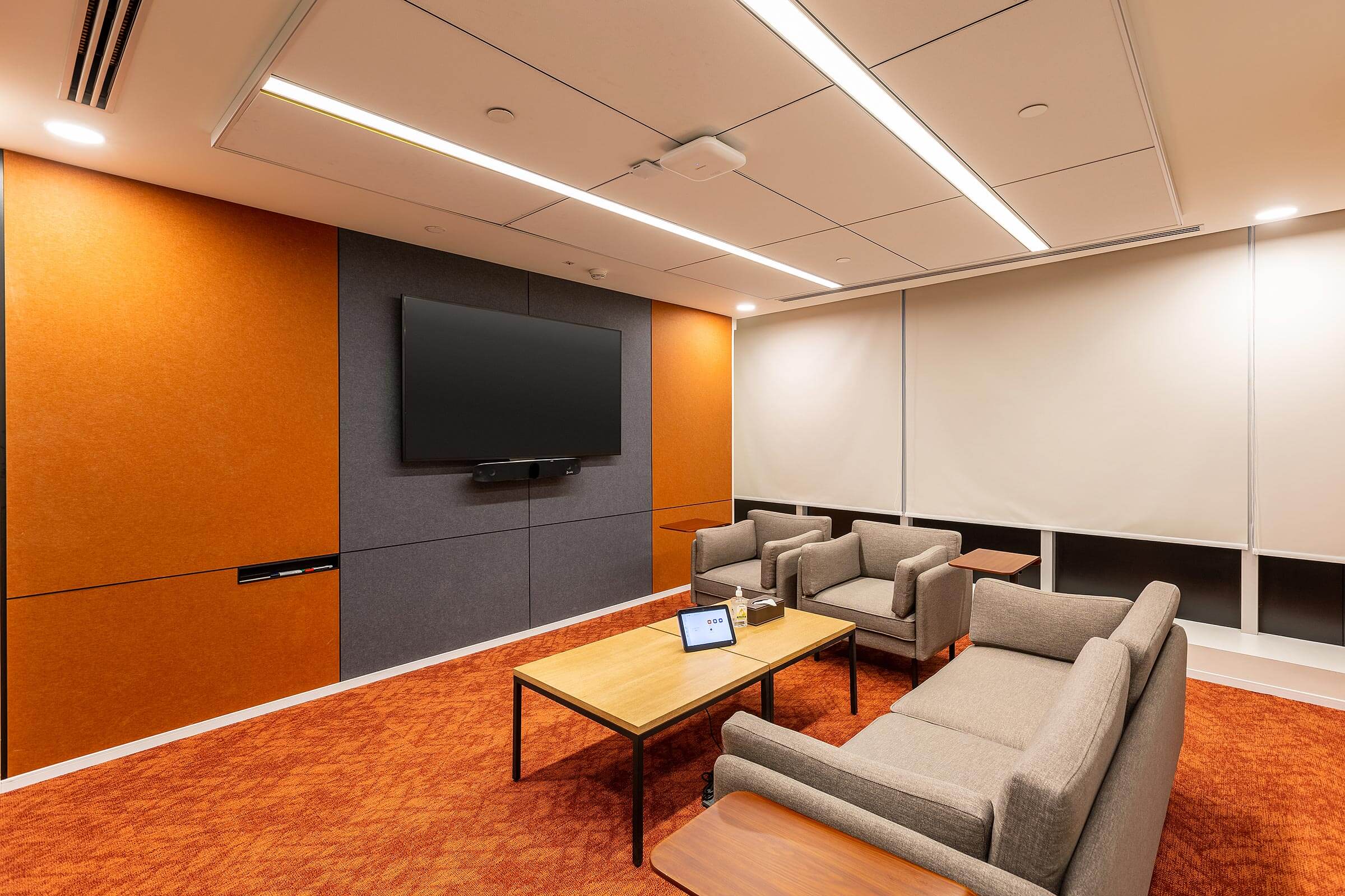 Compact meeting room with wall mounted screen, lounge seating, and orange carpet flooring